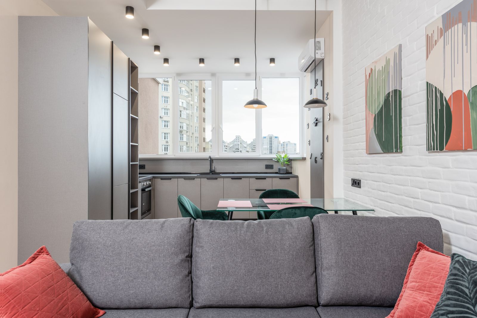 Gray sofa facing a dining table and kitchen in a city apartment, with art on a white brick wall